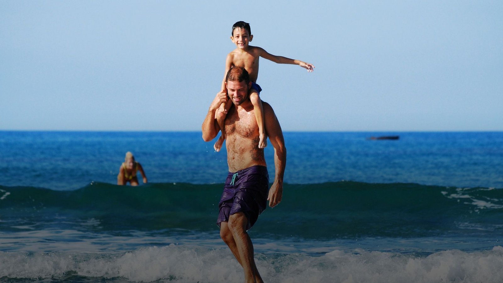 A father surfing a small wave with his son on his shoulders