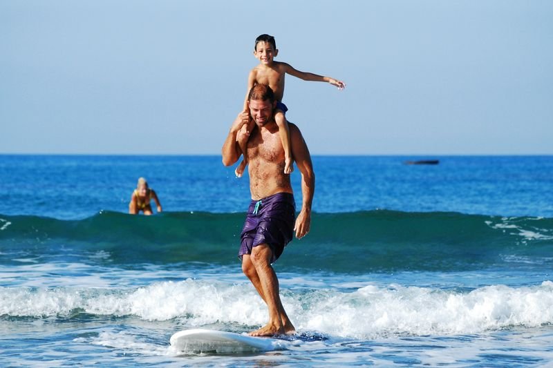 A father surfing a small wave with his son on his shoulders