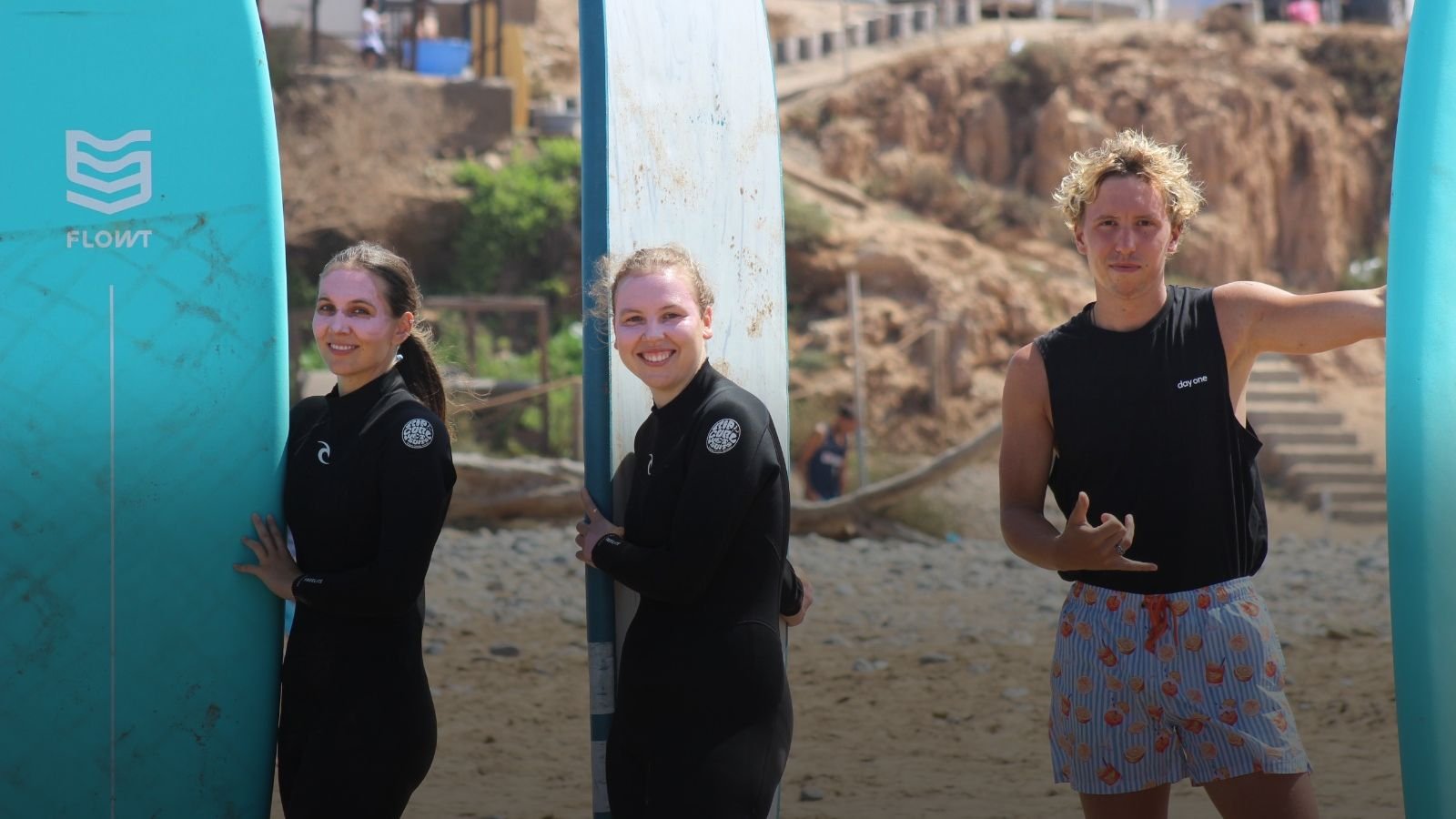 Drei Gäste mit ihren Surfboards am Strand: zwei lächelnde Frauen in schwarzen Neoprenanzügen halten lange Softboards aufrecht, ein junger Mann in schwarzem Tanktop und gemusterten Boardshorts zeigt das Shaka-Zeichen, dahinter eine Klippe und Stufen