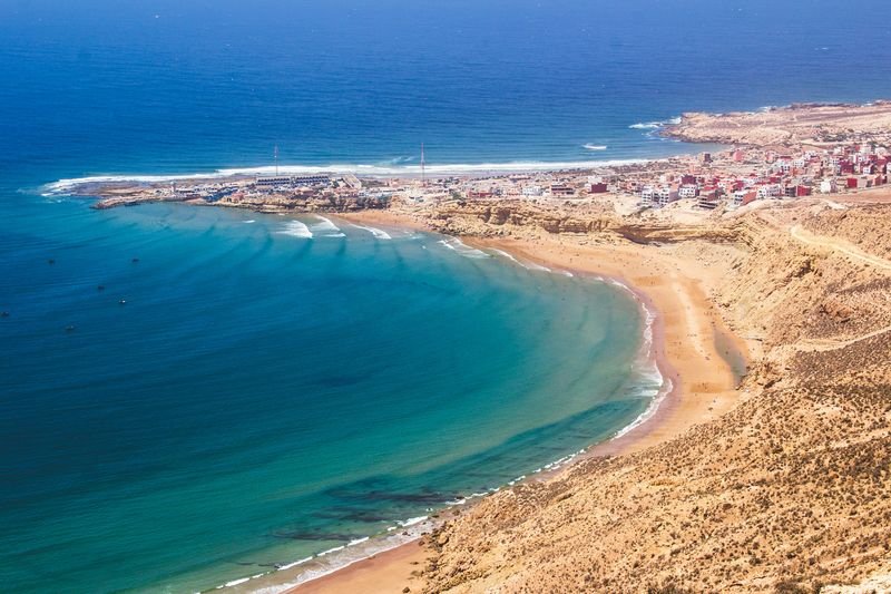Imsouane desde las colinas: líneas de swell que rodean la punta y entran en una bahía en curva, el pueblo y el espigón del puerto sobre el cabo, y una larga playa de arena al pie de laderas áridas