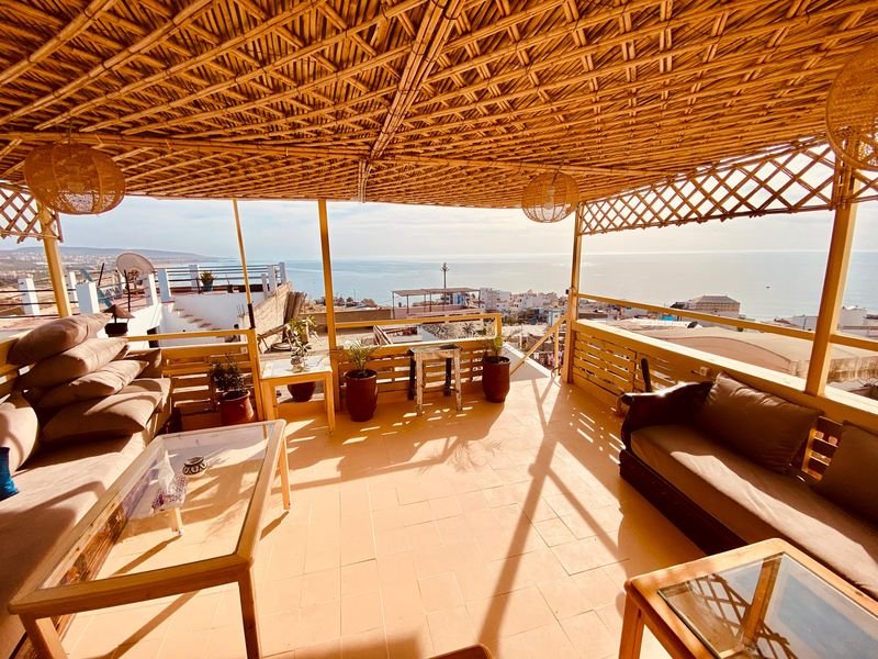 Dachterrasse des Hostels in Taghazout unter einer Schilfpergola: Sofas, ein niedriger Tisch und dahinter der Atlantik über dem Dorf