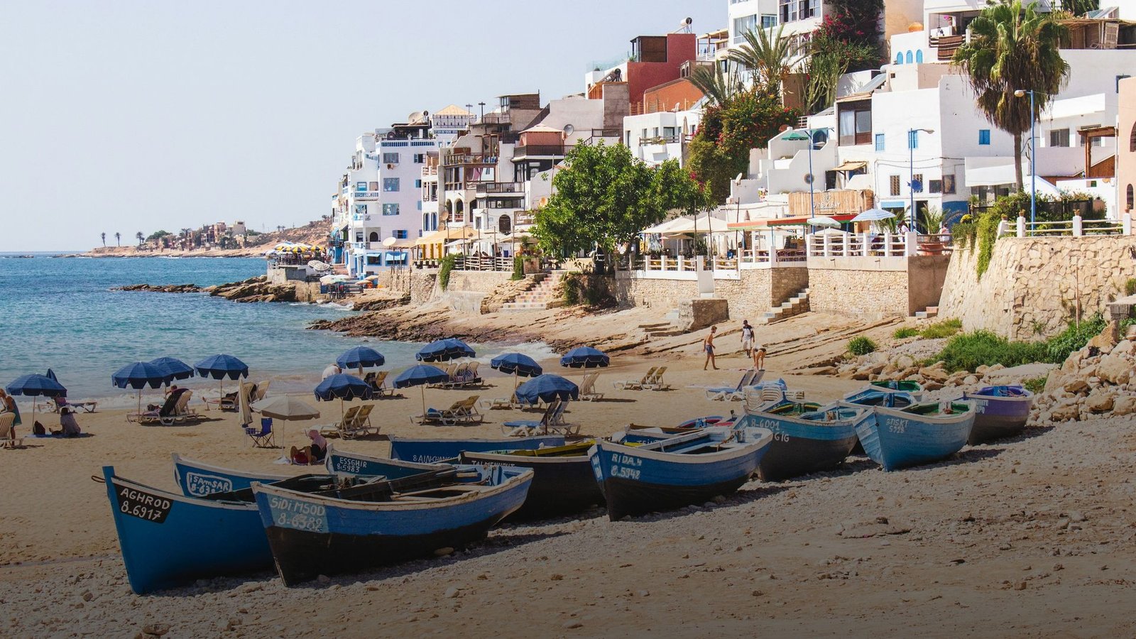 Playa de Taghazout: barcas azules de pescadores varadas en la arena, sombrillas y edificios blancos y de colores que suben por la ladera