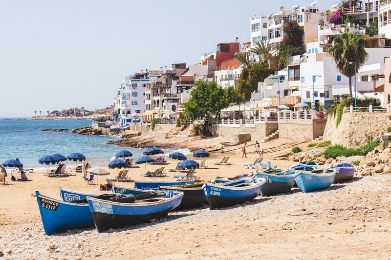 Taghazout beach: blue fishing boats hauled onto the sand, beach umbrellas, and white and coloured buildings climbing the slope behind