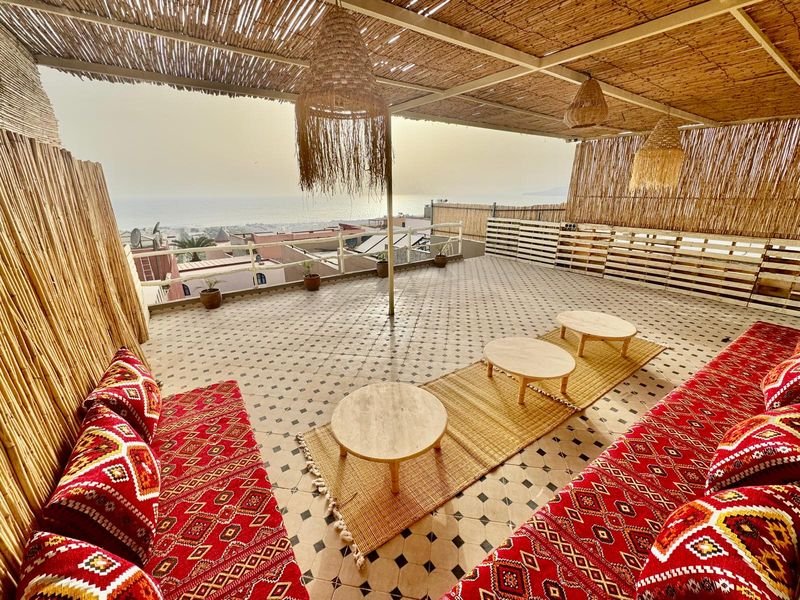 Dachterrasse des Hostels in Tamraght: rote Berberkissen, niedrige runde Tische und ein Sichtschutz aus Schilf, in der Ferne das Meer